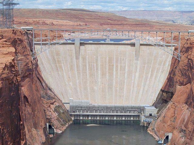 A front on photo of a massive dam built part way along a river. This giant man made structure in the Glen Canyon of USA holds back a large volume of water and is designed to remain strong for many years. 