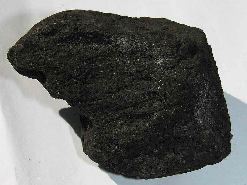 A close up photo that shows in detail the make up of a lump of coal. It's dark, sooty surface contrasts against the clean, light background. 