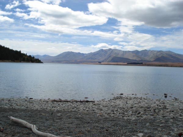 Looking out over the pebbles and rocks onto the flat surface of a lake. The calm conditions are helped by minimal wind and minimal could cover.