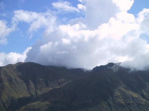 This photo shows a series of mountains which reach high into the air, piercing the base of the clouds and hiding their highest peak. 