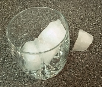 This photo shows a glass containing three ice cubes. There is also another ice cube sitting on the bench right next to the glass.