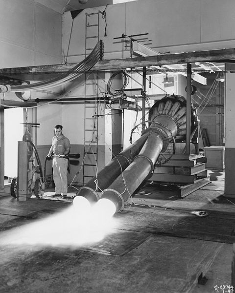 This is a black and white photo taken during a study of the effect of twin jet exhausts inclined toward the ground while in simulation of take-off conditions. A man stands on the left with a fire extinguisher, ready in the event of a fire.