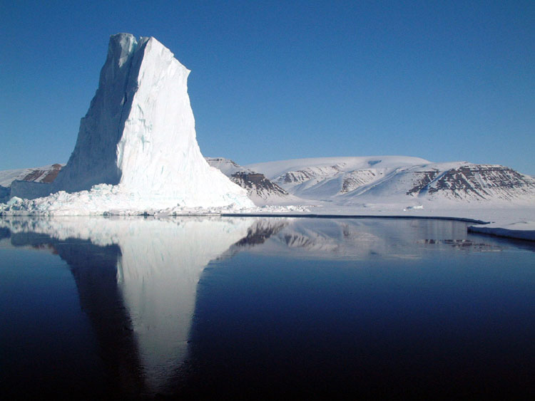 This beautiful image shows a unique looking iceberg in Greenland. There is a blue sky and the iceberg reflects off the cold water.