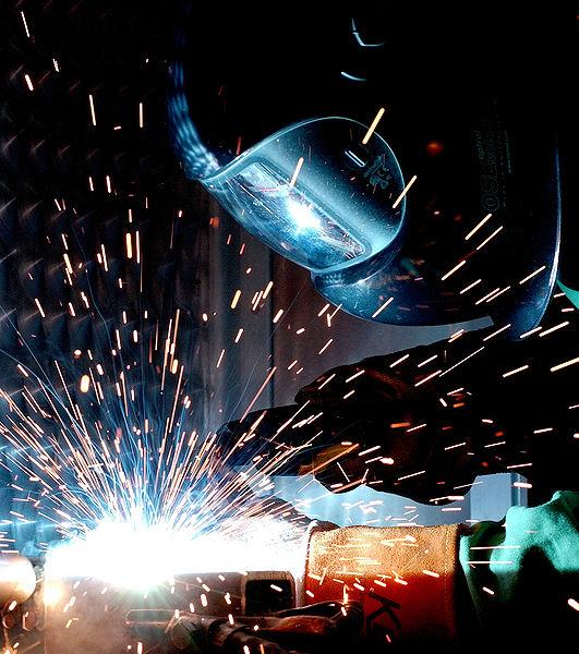 This photo shows a man metal arc welding. He is wearing important protective equipment such as a face mask and gloves. A large number of sparks fly off the metal as he works on the welding project.