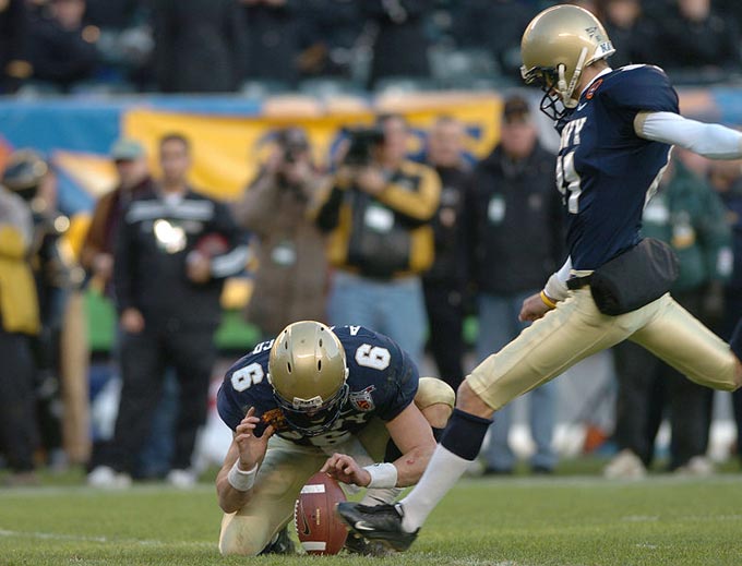 An American football kicker lines up his kick attempt as he makes his final stride before kicking the ball. His team mate holds the ball in position to help make the kick as accurate as possible.