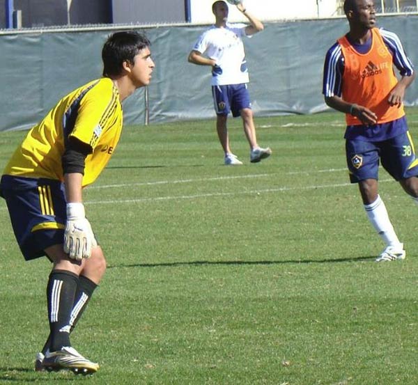 Two football (soccer) players take part in a training practice on a hot and sunny day while a trainer watches on in the image background.