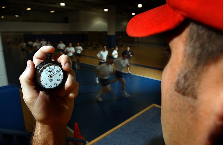 A trainer monitors the progress of a group as they run inside a gymnasium. The trainer is timing them using a stopwatch which can be seen in the image foreground.