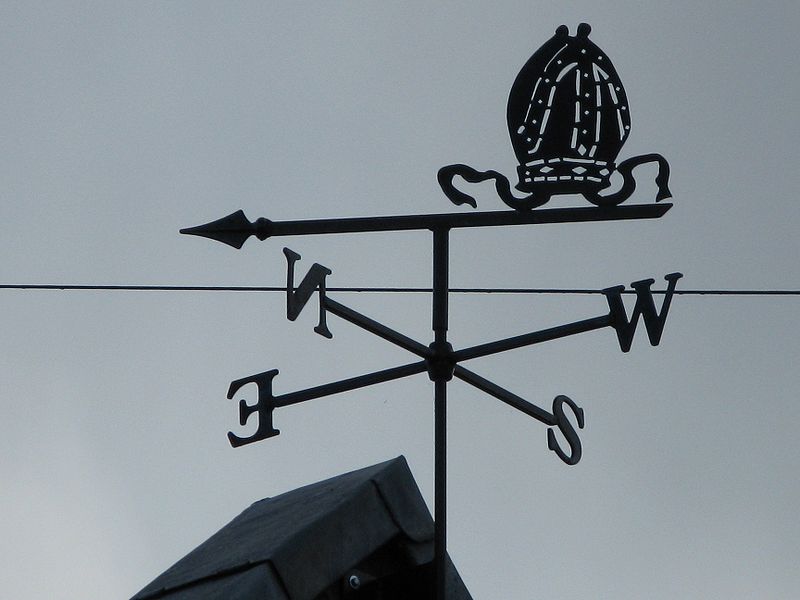 A weather vane can be seen on top of a house roof. It shows the letters N, S, E, W which represent north, south, east and west. It is used for wind direction.
